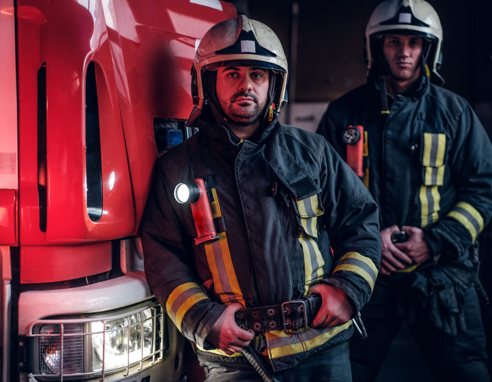 Two firemen standing near the fire truck at night in a fire depot