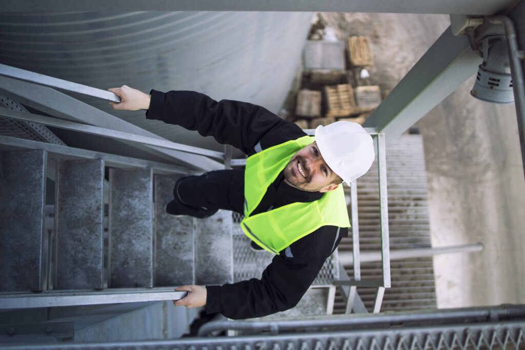 Smiling industrial worker wearing PPE climbs a metal staircase on a silo structure.